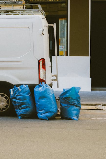 The image depicts the rear side of a white delivery van parked on a street next to a building. Adjacent to the van, three large blue rubbish bags made of plastic are positioned on the pavement, partia